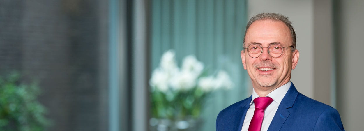 Persoon in formele kleding met bril staat binnen in een lichte ruimte, met een raam en een vaas met witte bloemen op de achtergrond.